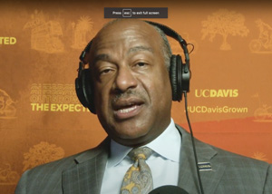 Close up of Chancellor Gary S. May talking with head phones on in front of an orange background